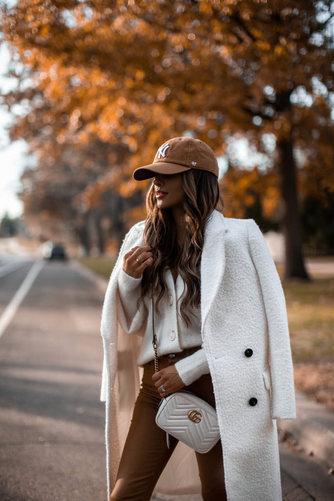 fashion blogger mia mia mine wearing a white coat from express with a cardigan and ny yankees baseball cap
