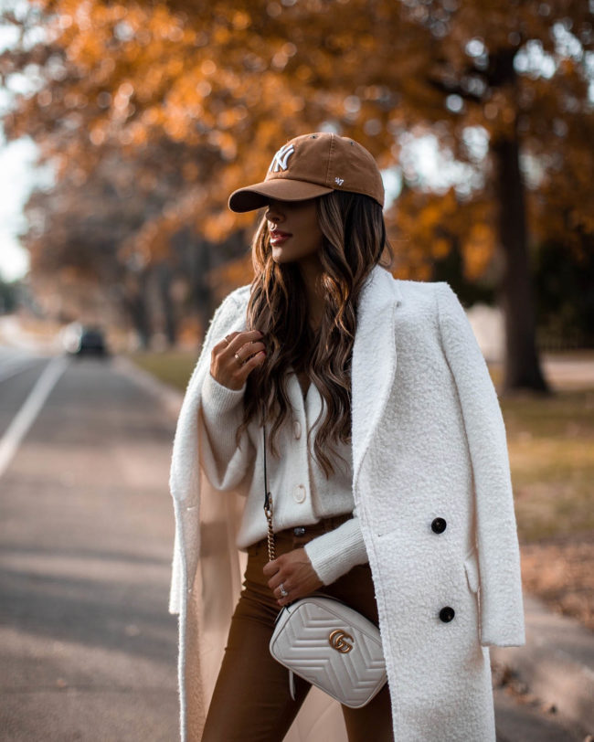 fashion blogger mia mia mine wearing a white coat from express with a cardigan and ny yankees baseball cap